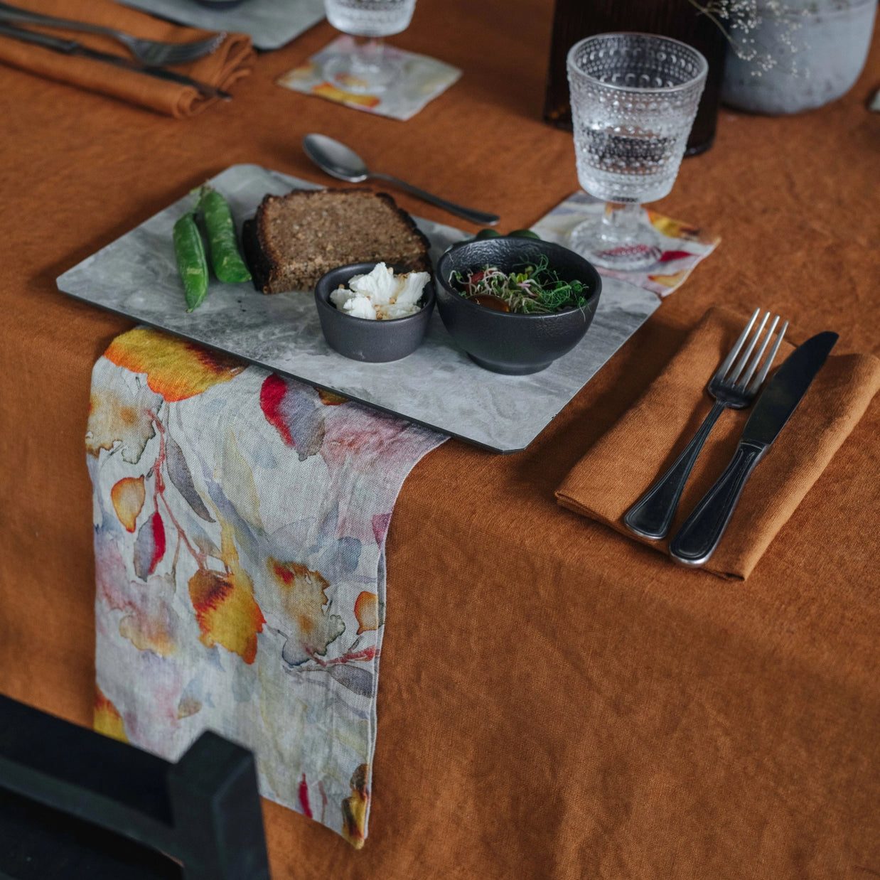 Slate serving board with dark bread, snap peas, goat cheese, and microgreens on rust velvet tablecloth with crystal glass