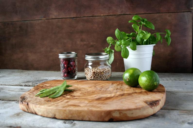 Round olive wood cutting board with natural edge grain on weathered white surface with fresh limes and sage leaves