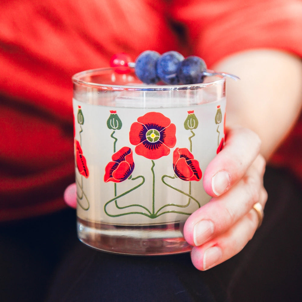 Clear glass tumbler with red poppy flower design and green stems, garnished with three blueberries on cocktail pick