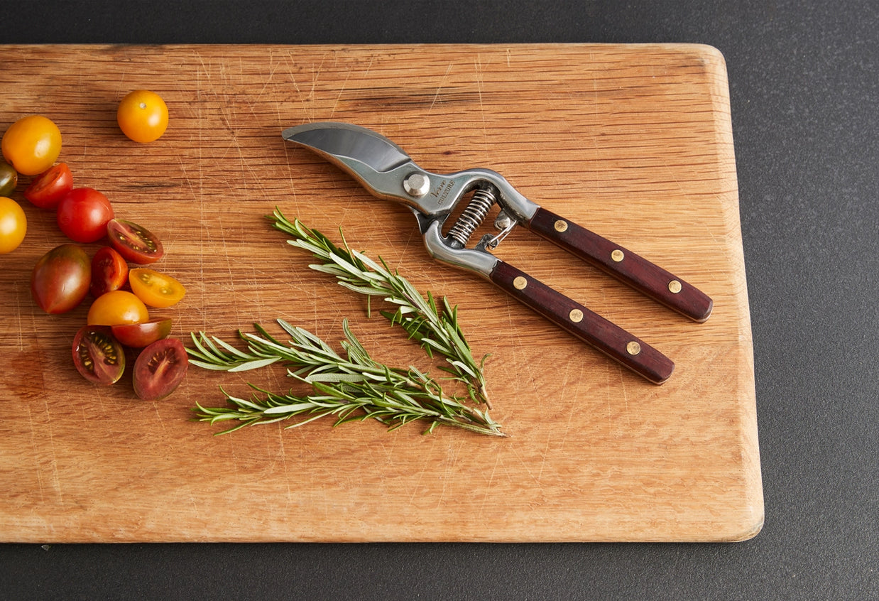 Garden shears with dark wood handles and brass rivets on oak cutting board with cherry tomatoes and fresh rosemary sprigs
