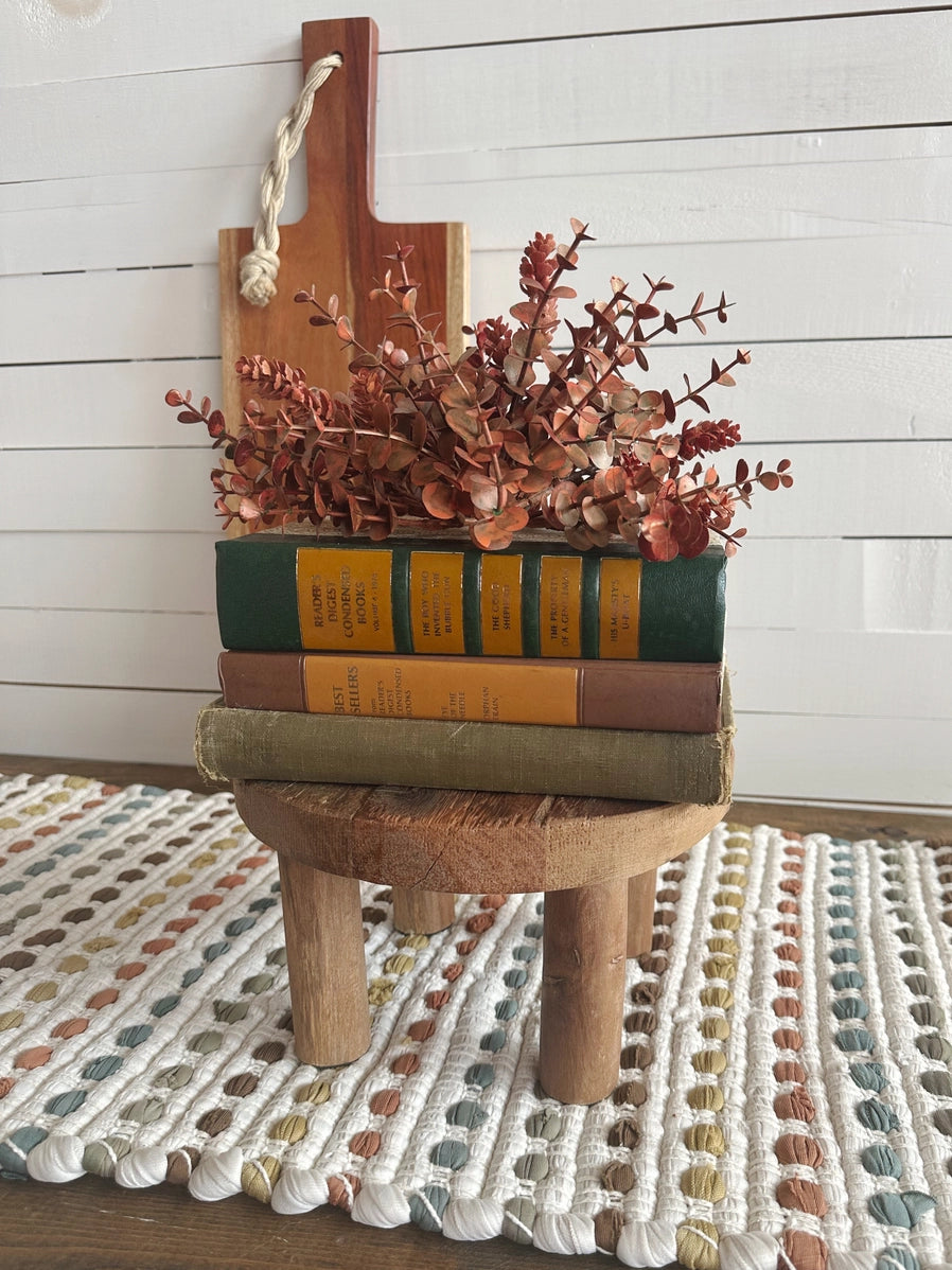 Farmhouse vignette with dried rust eucalyptus, vintage green and tan books stacked on round wooden stool with rope handle