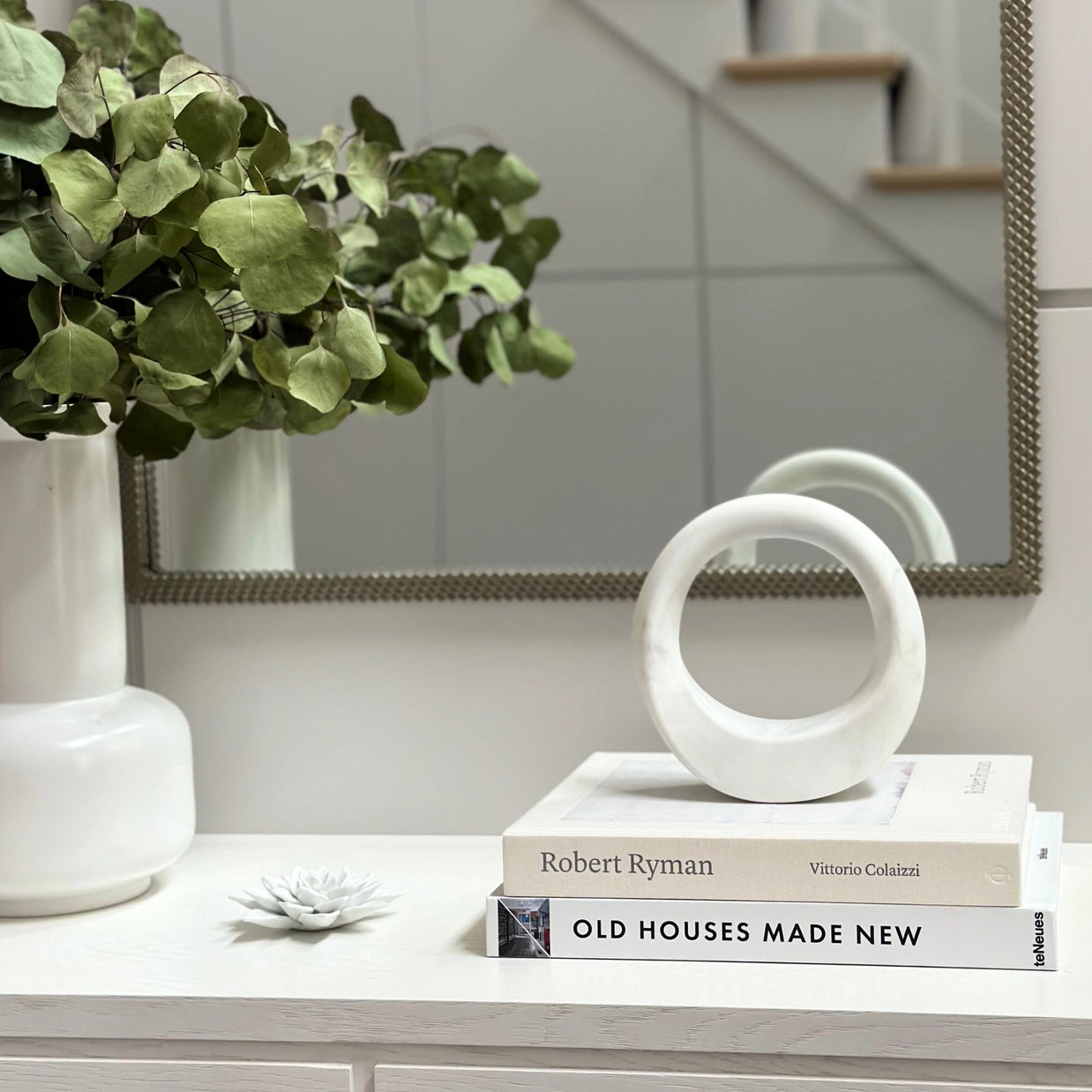 White ceramic sculptural object with circular cutout rings stacked on coffee table books next to eucalyptus arrangement