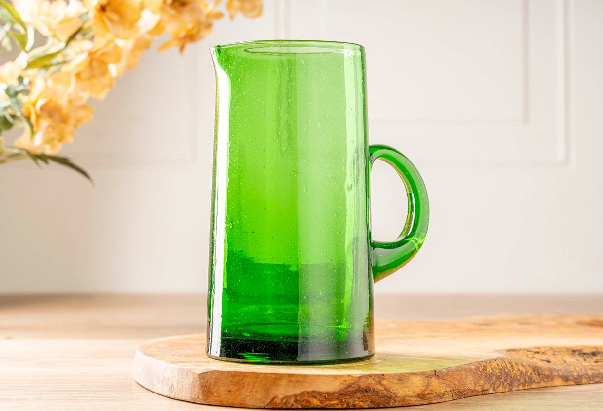Bright lime green glass pitcher with round handle and tapered cylindrical shape on natural wood serving board