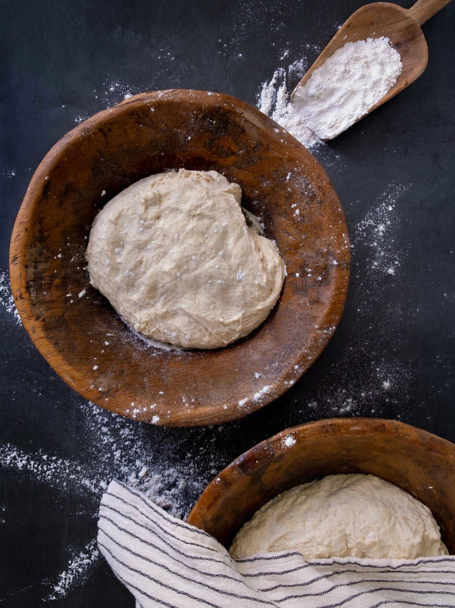 Raw bread dough balls in wooden bowls with flour-dusted wooden scoop and striped linen towel on dark surface
