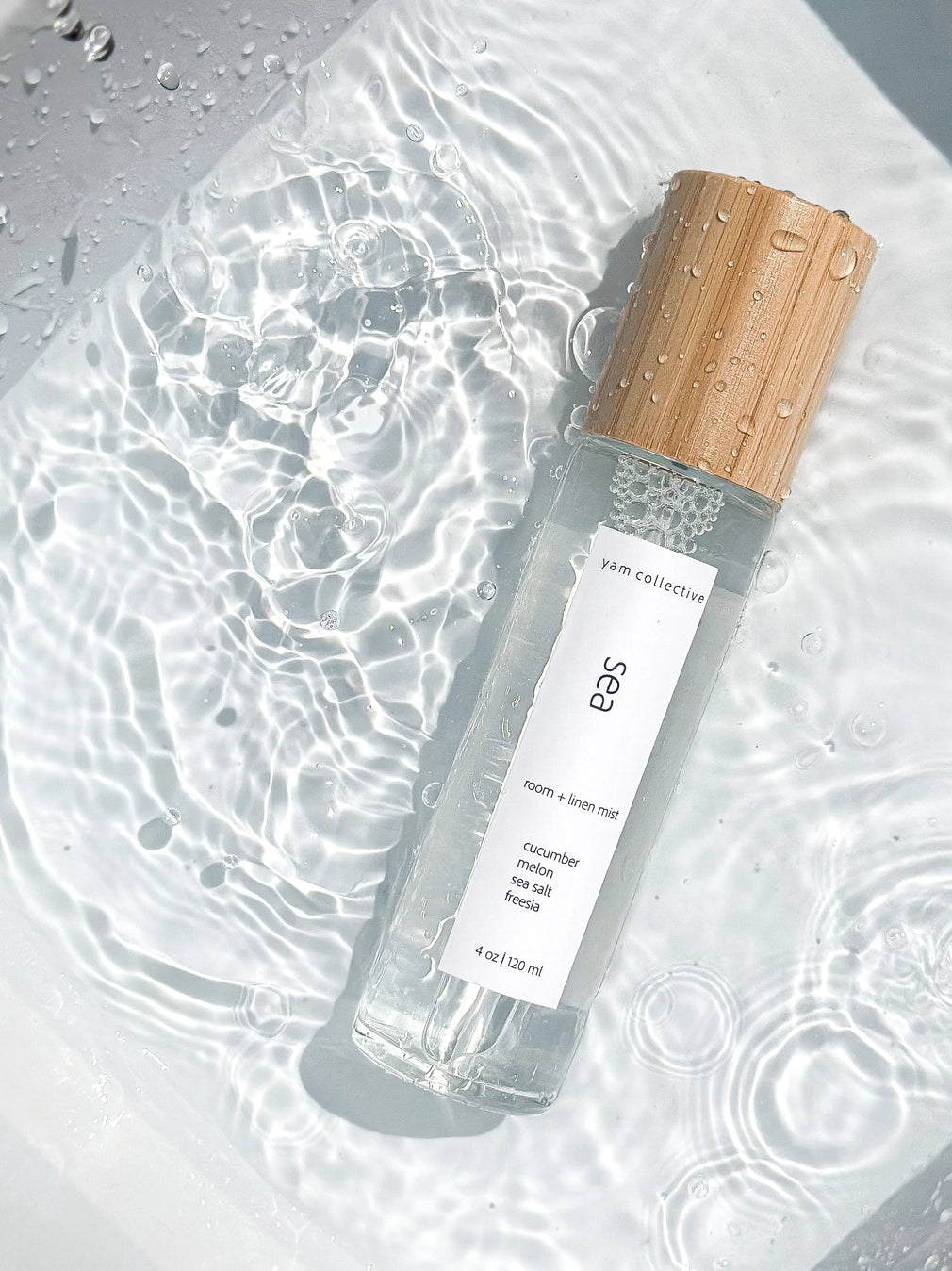 Clear glass room spray bottle with bamboo wood cap and white label, covered in water droplets on wet surface