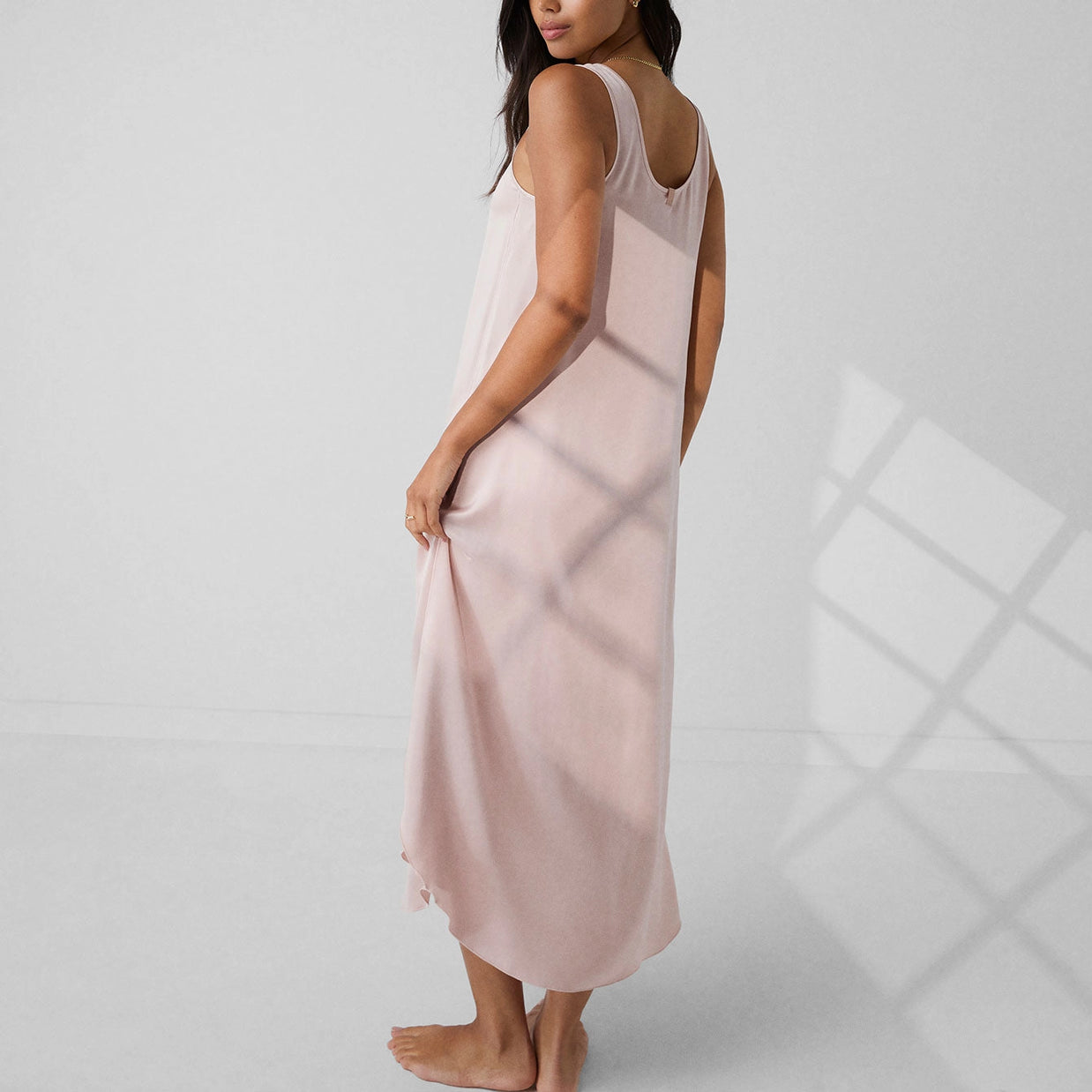 Blush pink satin slip dress with scoop back and asymmetrical hem worn by model shown from behind on light background