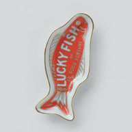 A fish-shaped ceramic dish or tray with a red center, white outer area, gold trim, and the words 'LUCKY FISH or GOOD FORTUNE' printed on it.