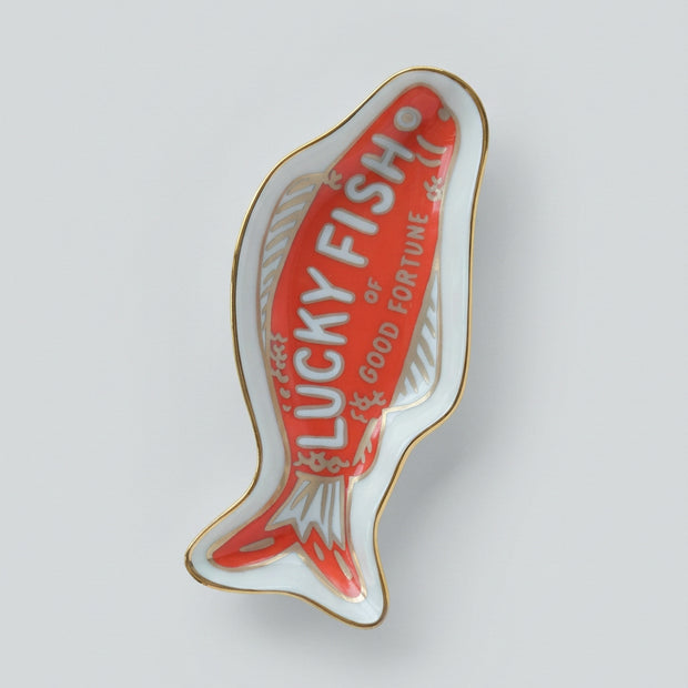 A fish-shaped ceramic dish or tray with a red center, white outer area, gold trim, and the words 'LUCKY FISH or GOOD FORTUNE' printed on it.