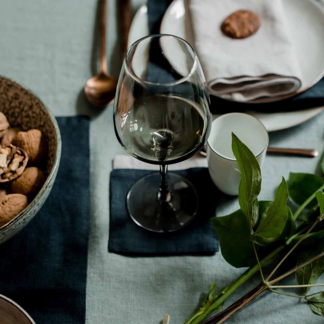 Smoke gray stemmed wine glass with tapered bowl on navy linen runner beside copper serving utensils and white candle