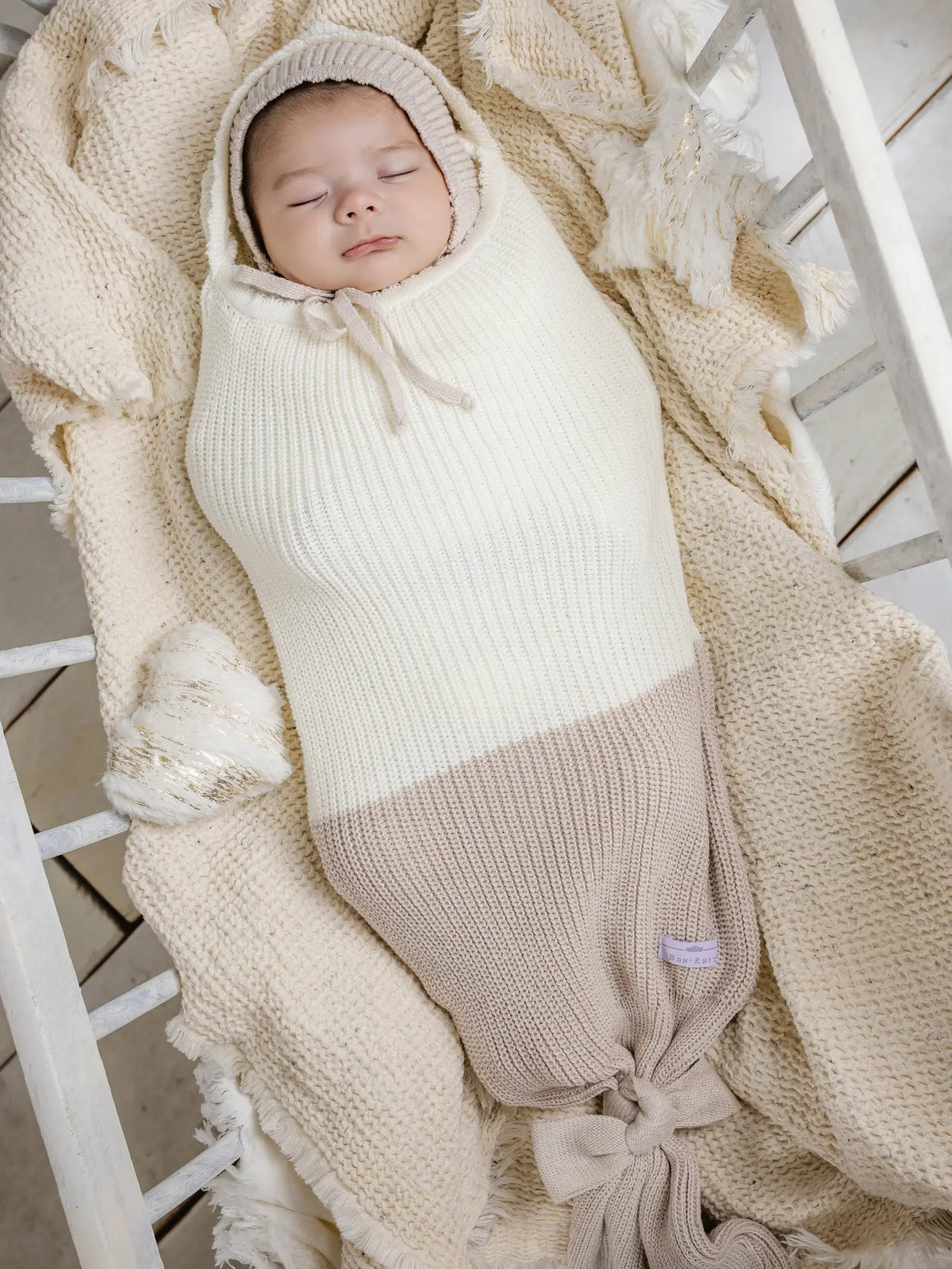 Cream and taupe knit baby cocoon swaddle with matching bonnet, featuring ribbed texture and knotted bottom closure