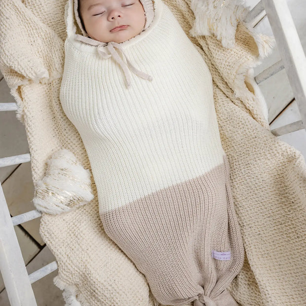 Cream and taupe knit baby cocoon swaddle with matching bonnet, featuring ribbed texture and knotted bottom closure