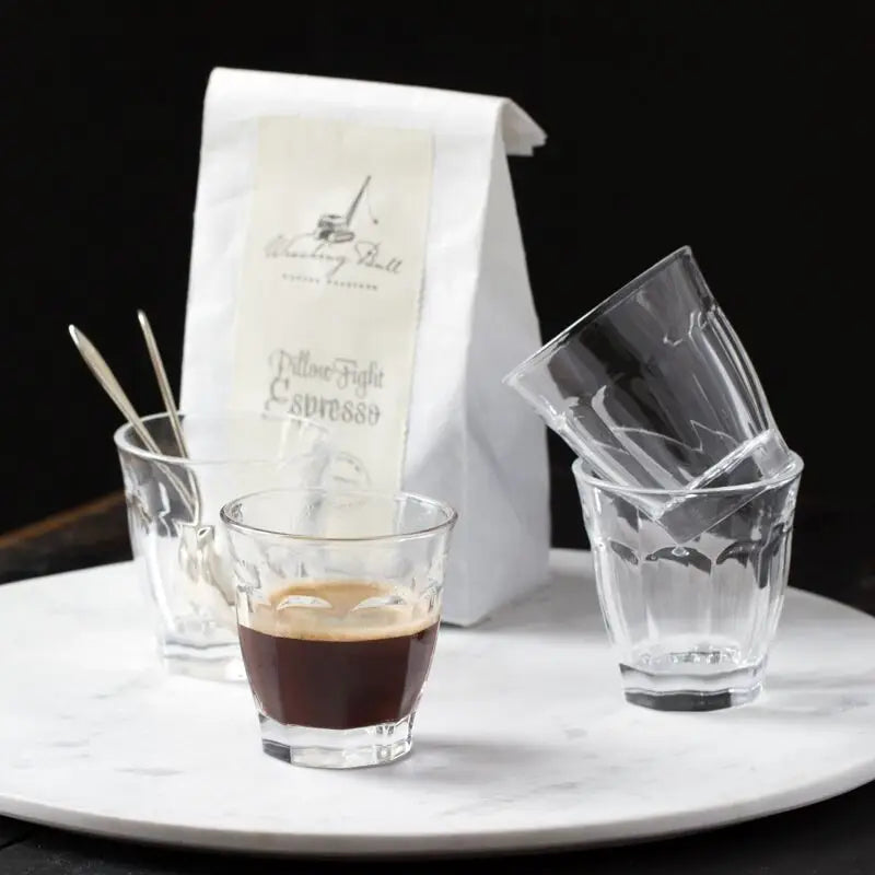 Espresso shot in faceted clear glass tumbler with white coffee bag and stacked empty glasses on marble serving plate