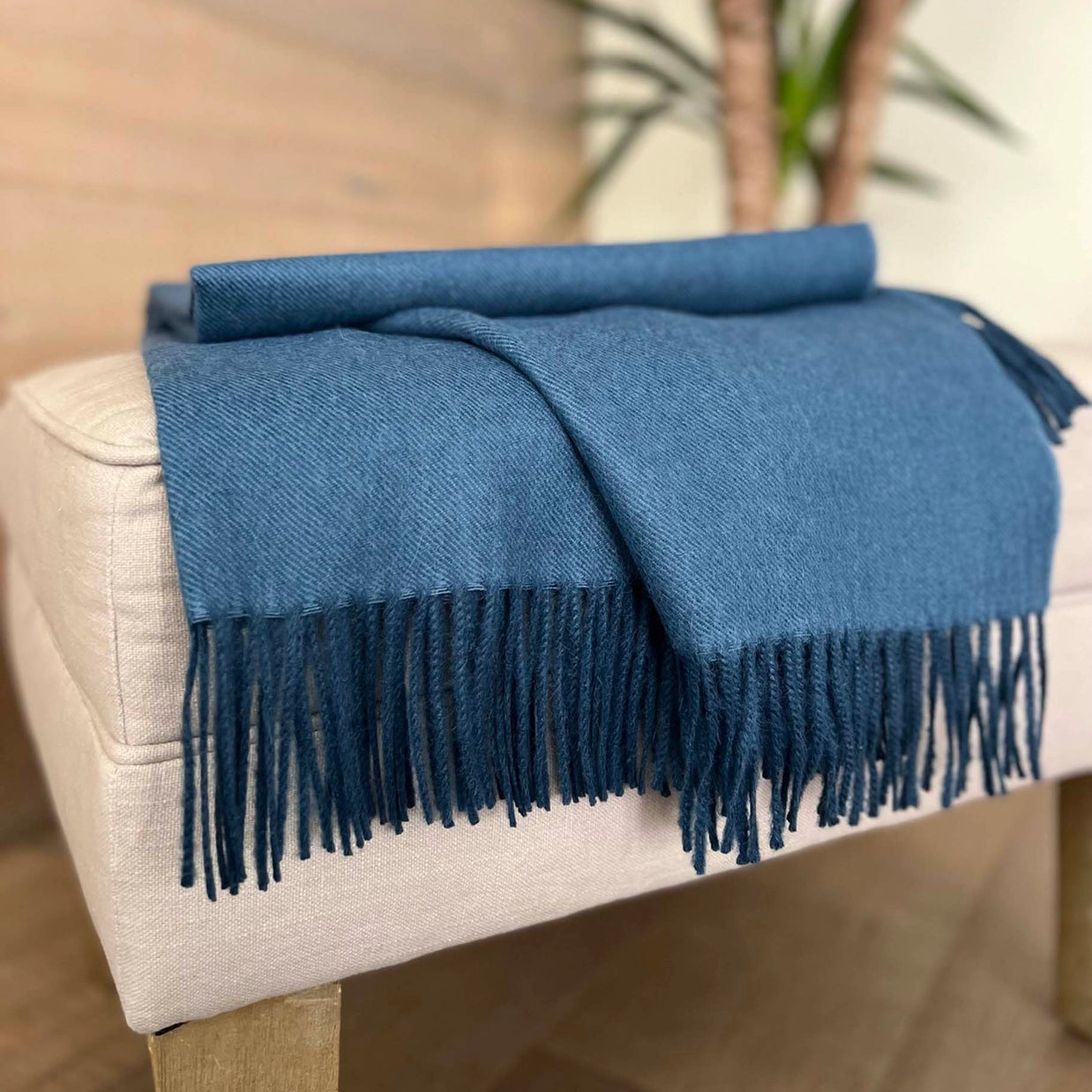 Navy blue woven throw blanket with diagonal twill texture and long knotted fringe draped over beige upholstered ottoman