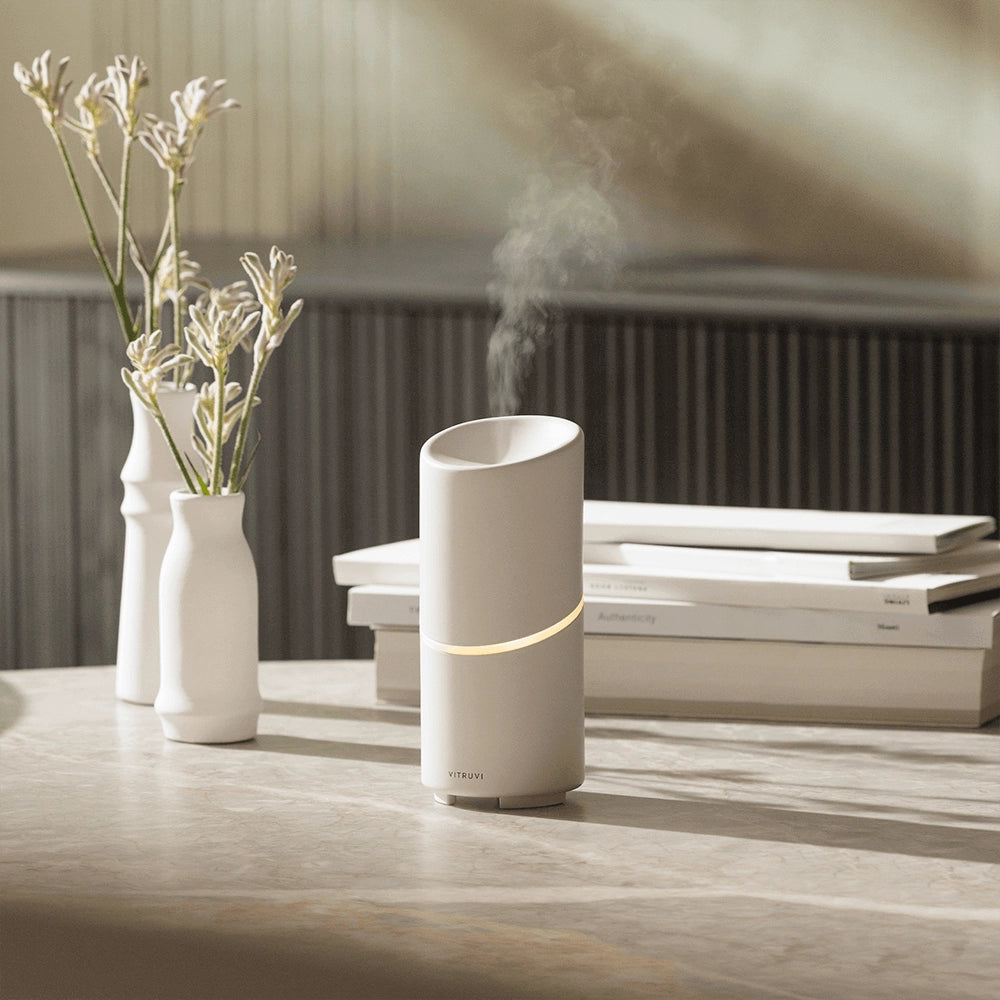 Matte white cylindrical aromatherapy diffuser with gold band accent releasing mist on marble table with botanical decor
