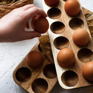 Wooden egg holder tray with circular indents holding brown and tan eggs, hand placing egg in natural wood organizer