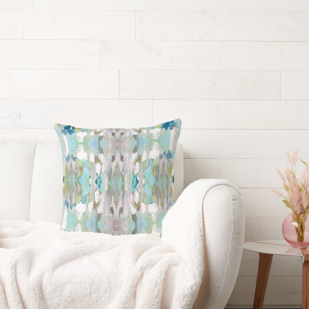Throw pillow with abstract watercolor pattern in turquoise, sage green, gray, and white on cream fabric with self-piping