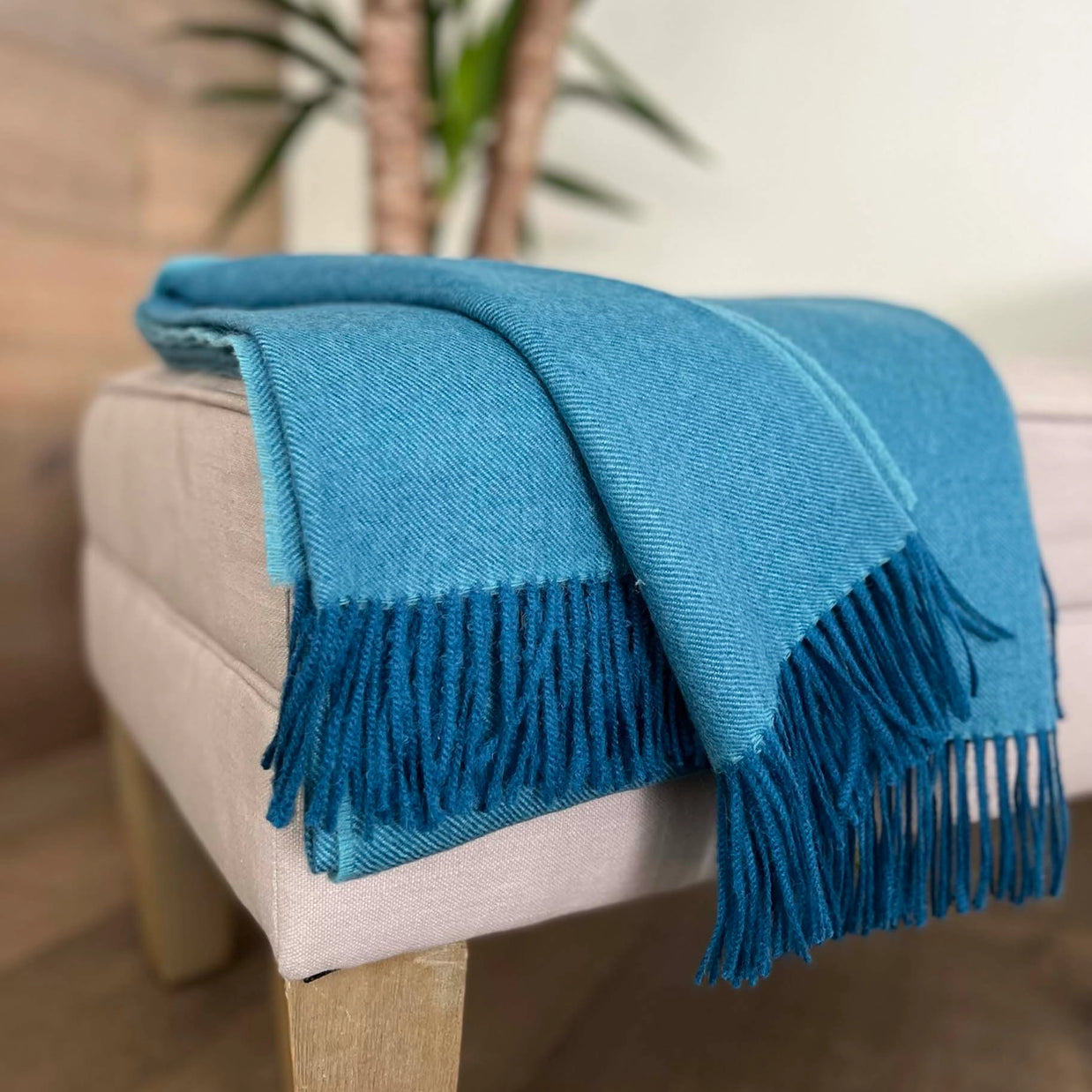 Teal blue woven throw blanket with diagonal twill texture and navy fringe trim draped over beige linen ottoman