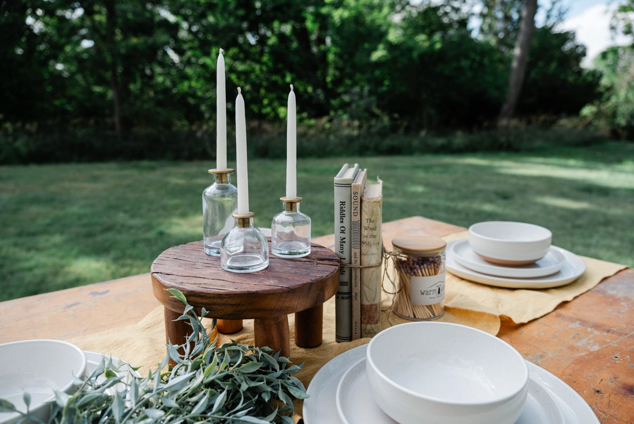 Three clear glass bottle candle holders with brass collars holding white taper candles on wooden pedestal stand