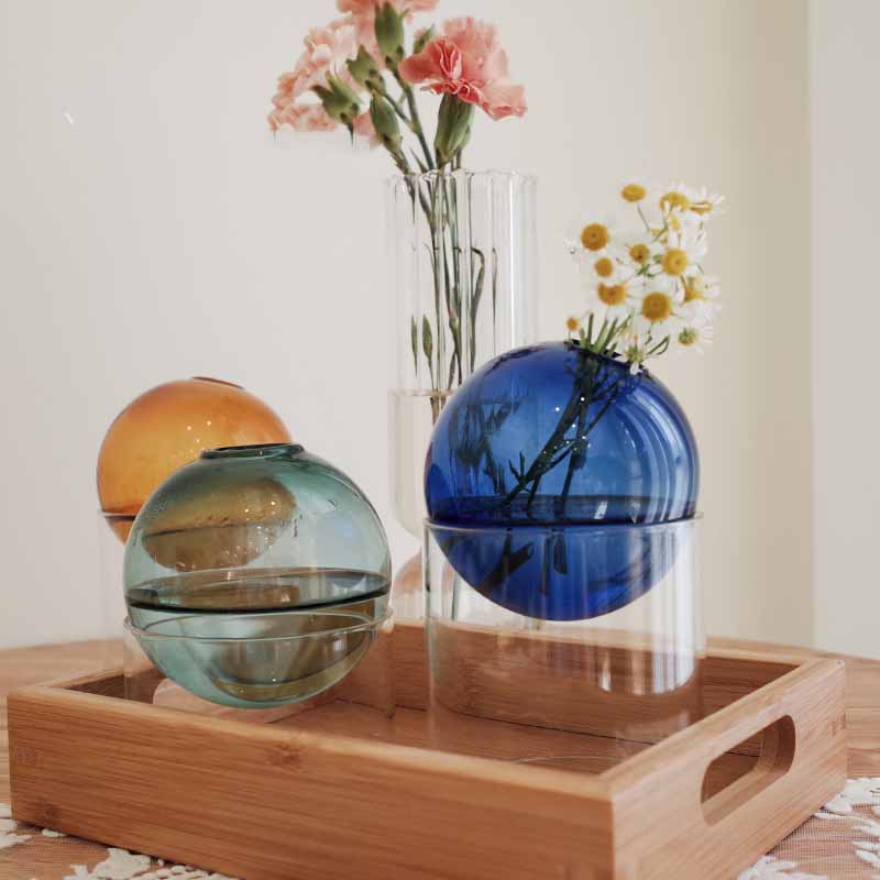 Spherical glass vases in blue and teal with clear stands on wooden tray, holding pink carnations and white daisies