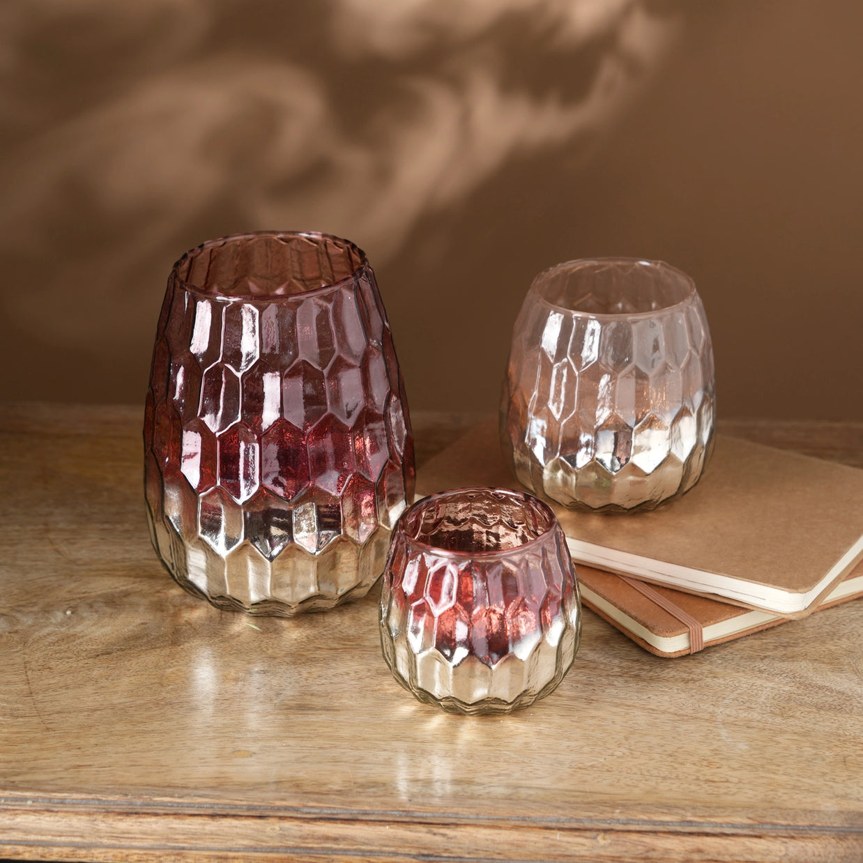 Three faceted glass votive holders with honeycomb pattern in rose gold ombre finish on wooden surface with notebook
