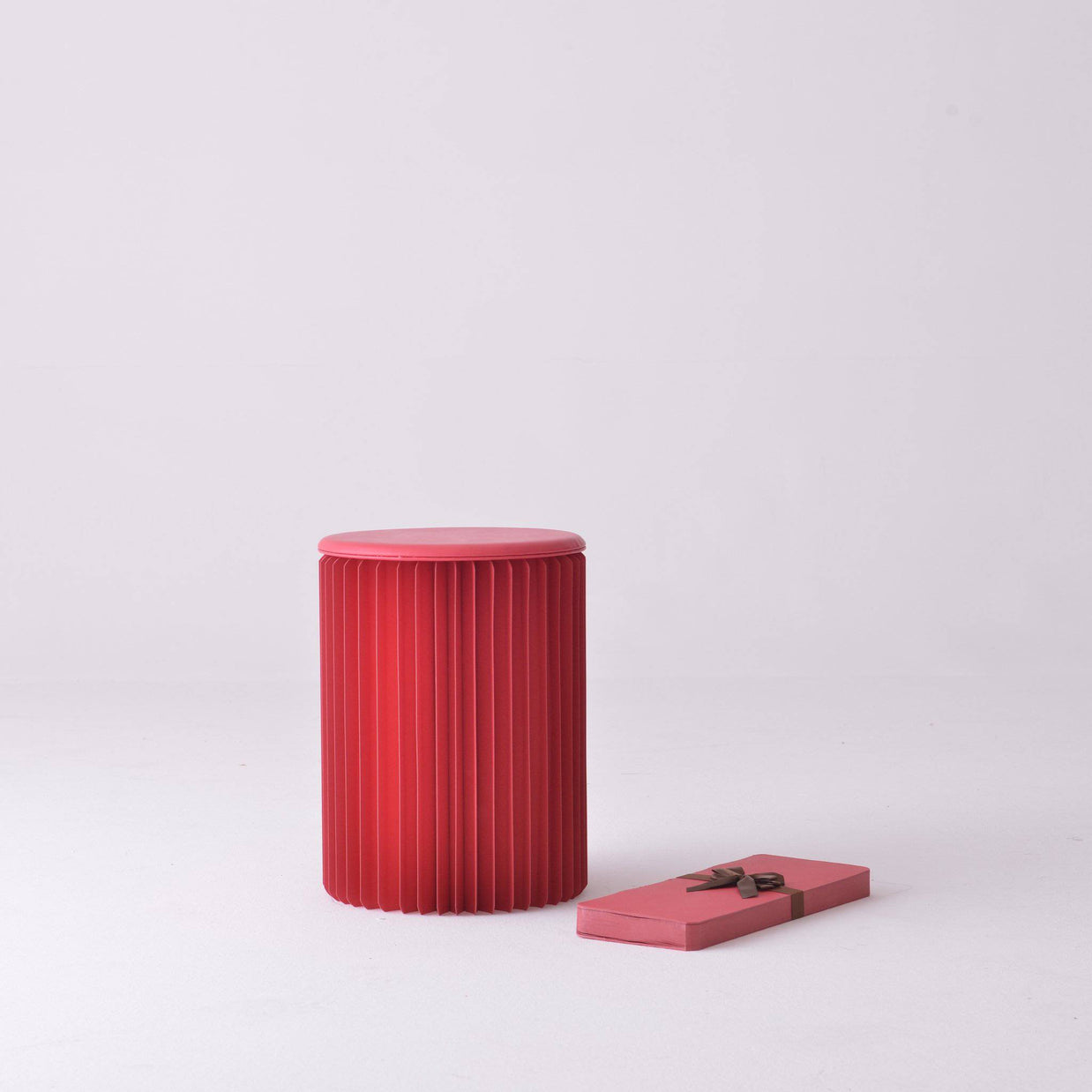 Red cylindrical stool with vertical pleated ridges and smooth rounded top, shown with decorative gift box