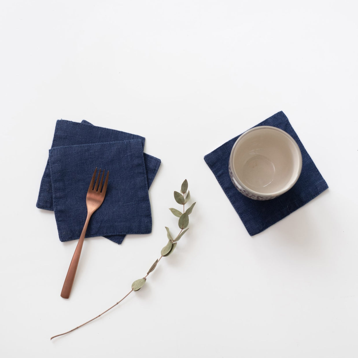 Navy blue linen coasters with textured weave, displayed with copper fork and cream ceramic bowl on white surface