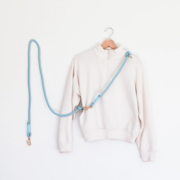 A blue hands-free braided rope dog leash hanging on a wooden hanger with a light gold hardware, displayed alongside a light-colored sweater.