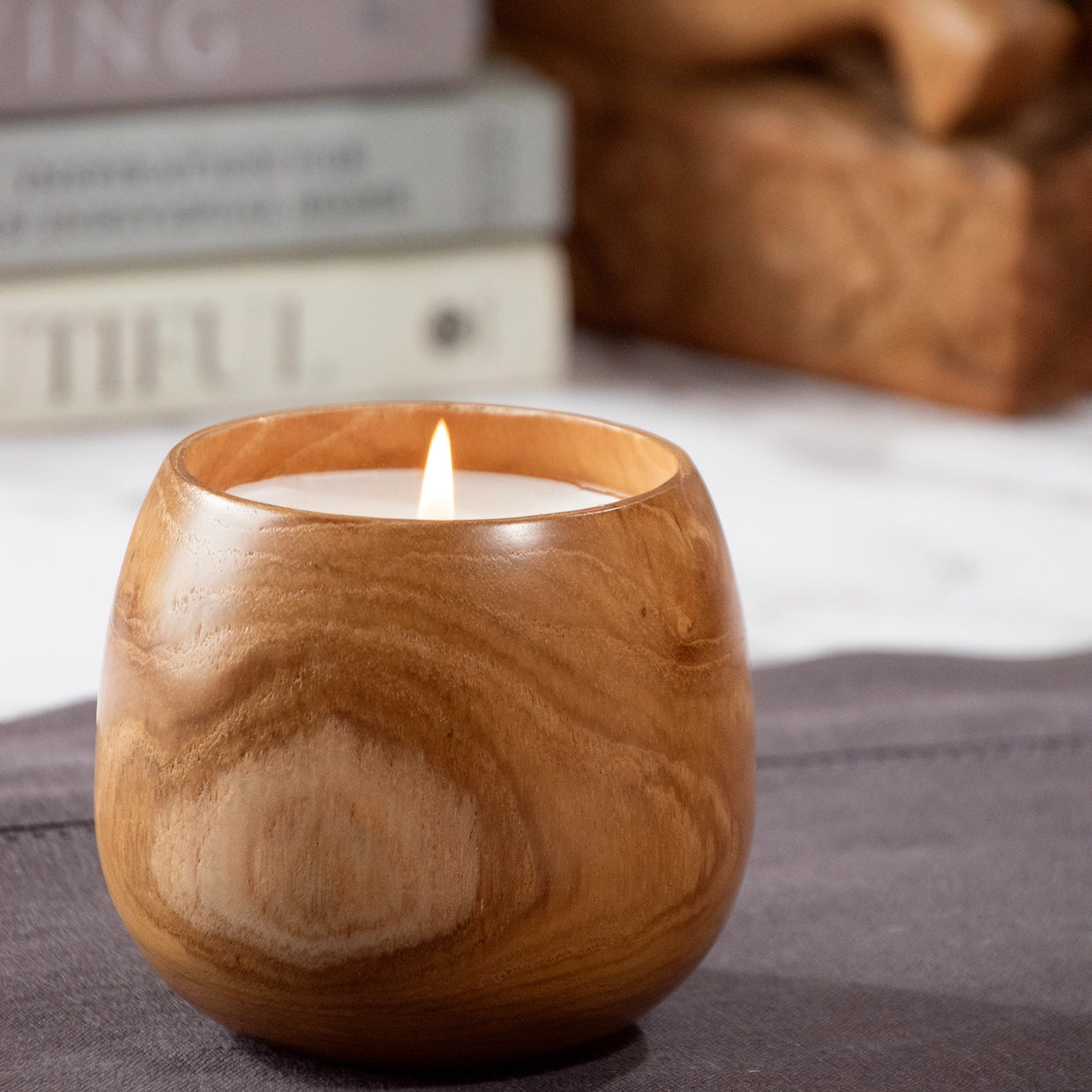 Round wooden bowl candle with natural grain pattern, cream wax, and lit flame on gray surface with blurred background