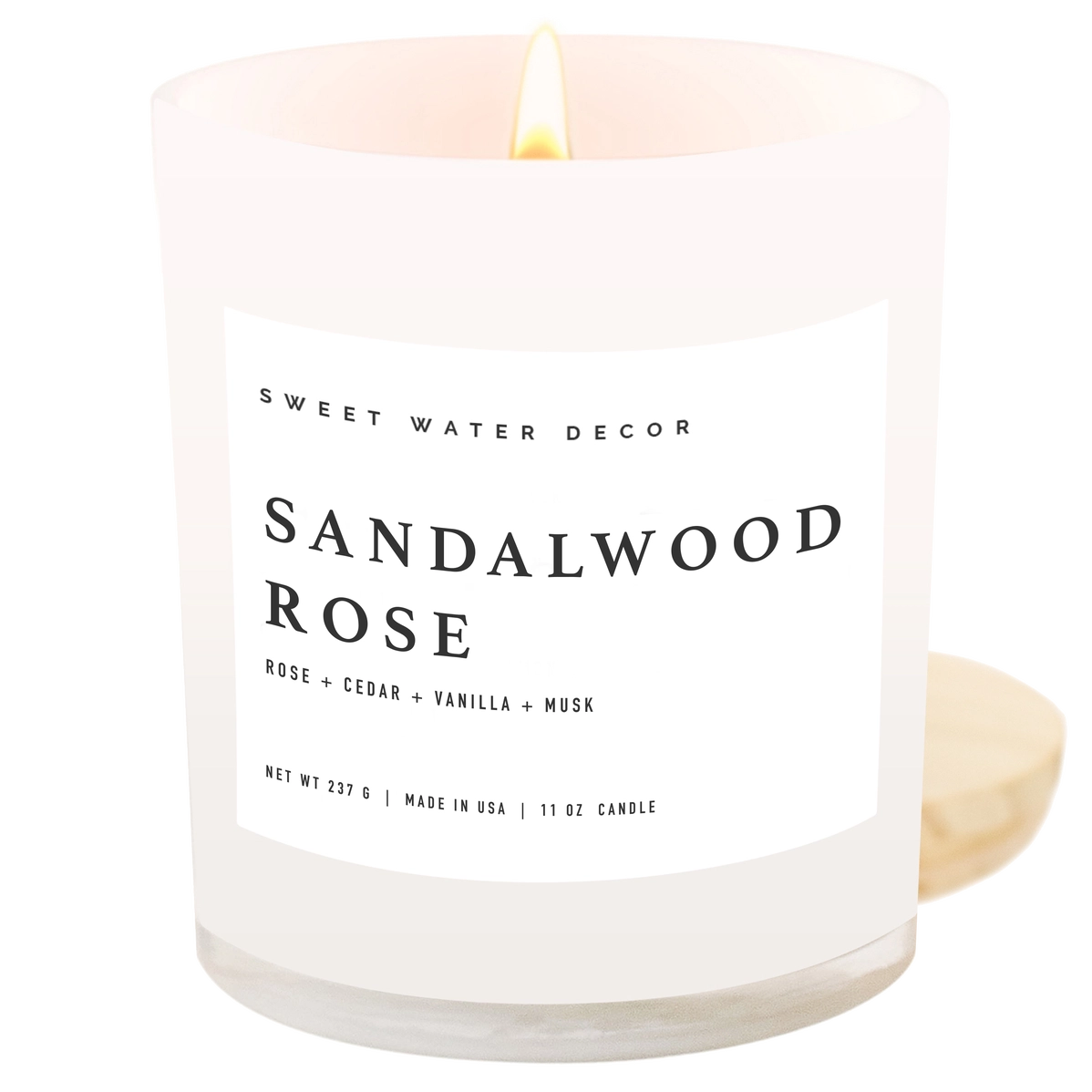 Frosted white glass candle with lit flame, minimalist black label reading Sandalwood Rose with rose, cedar, vanilla, musk notes