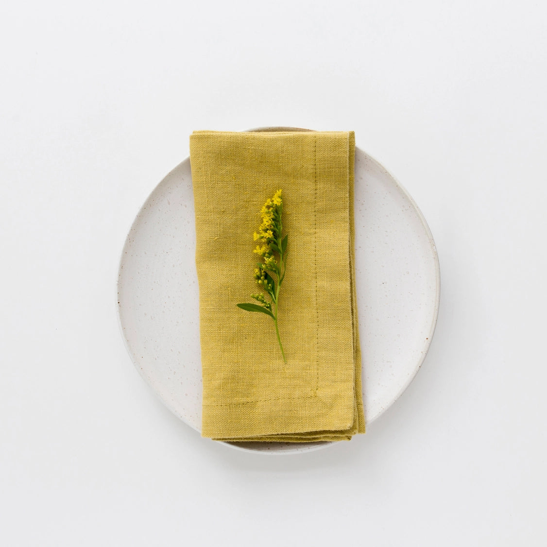 Golden yellow linen napkin folded on white speckled ceramic plate, topped with yellow wildflower sprig