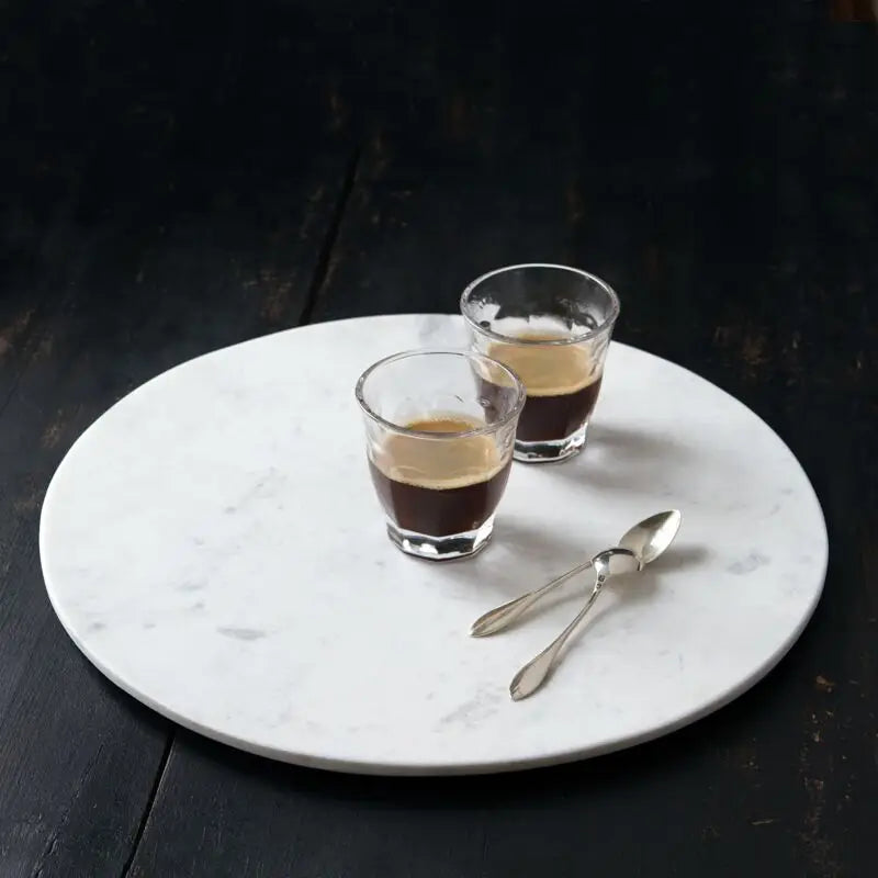 Round white marble serving board with gray veining, displayed with two espresso glasses and silver spoons on dark surface