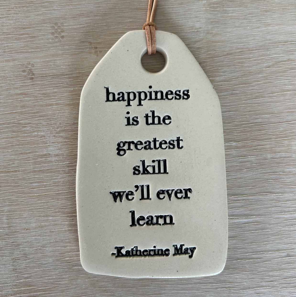 White ceramic tag with black text quote on leather cord hanging against weathered gray wood surface