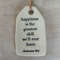 White ceramic tag with black text quote on leather cord hanging against weathered gray wood surface