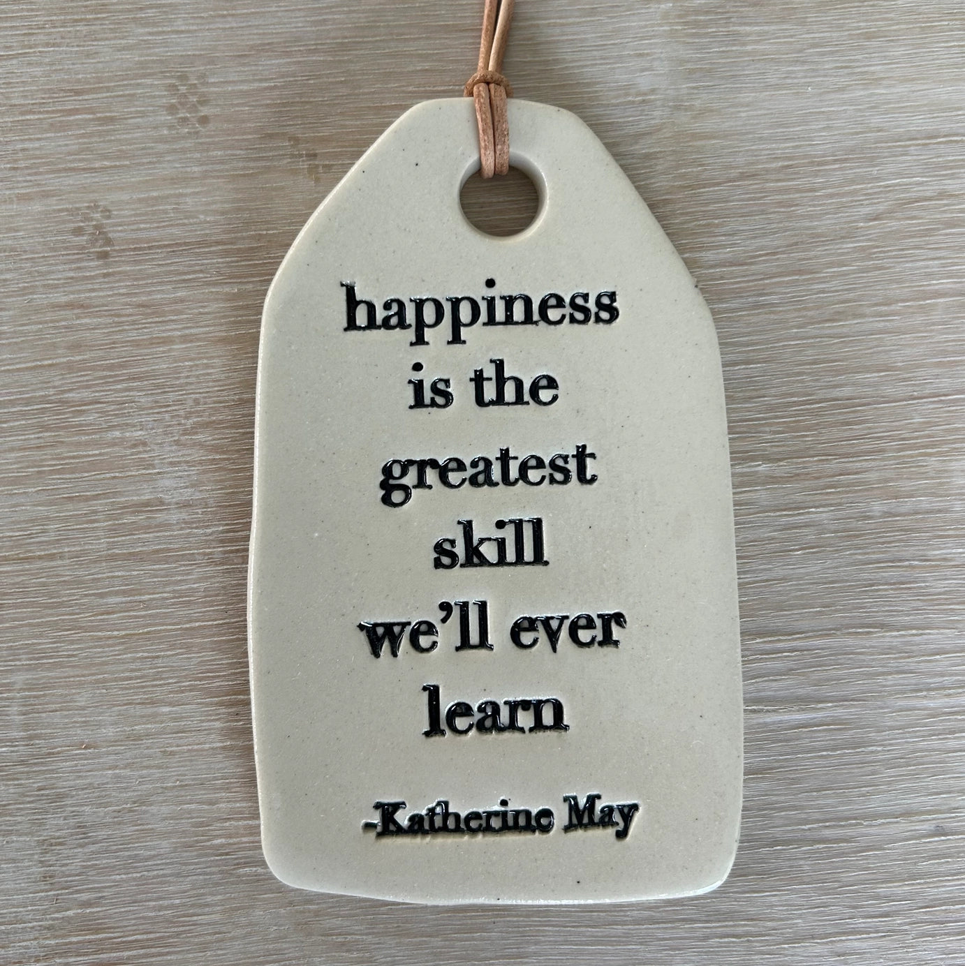 White ceramic tag with black text quote on leather cord hanging against weathered gray wood surface