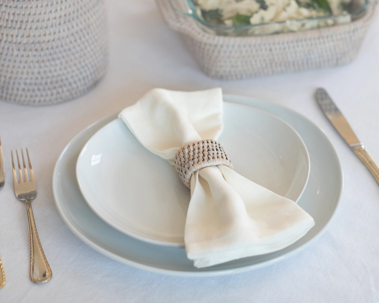 Woven rattan napkin ring holding crisp white linen napkin on pale blue ceramic dinnerware with gold flatware