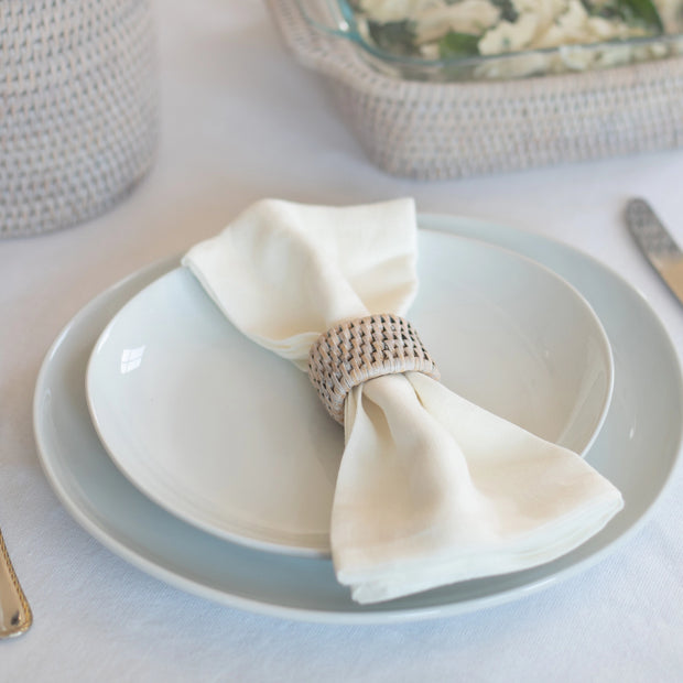 Woven rattan napkin ring holding crisp white linen napkin on pale blue ceramic dinnerware with gold flatware