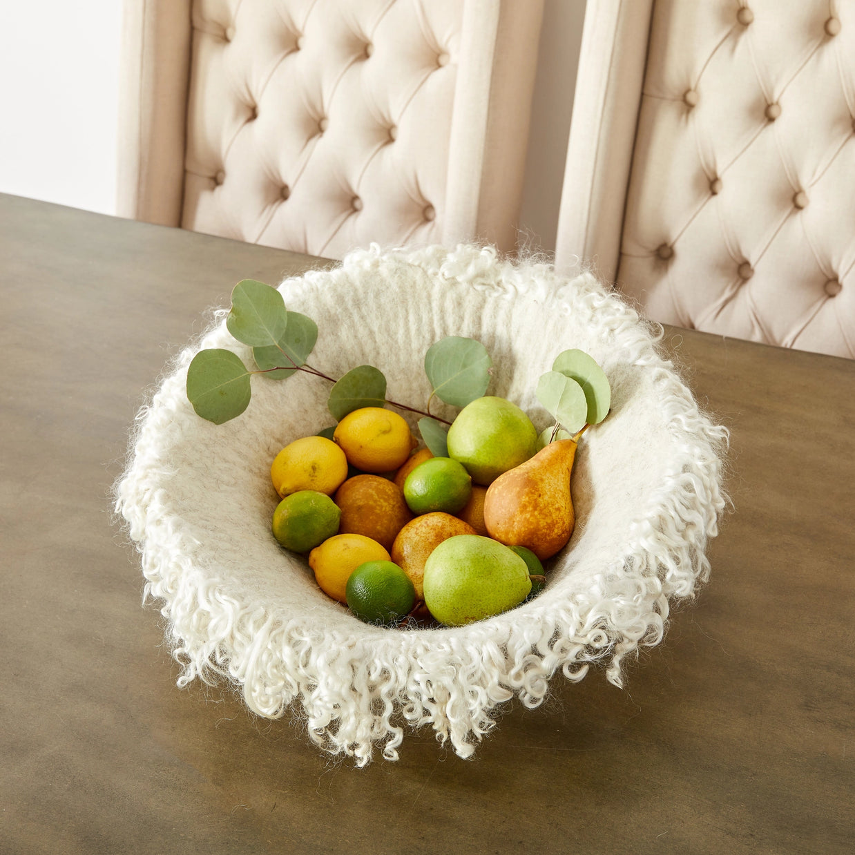 Cream wool basket with curly fringe edges filled with citrus fruits, pears, and eucalyptus on gray dining table