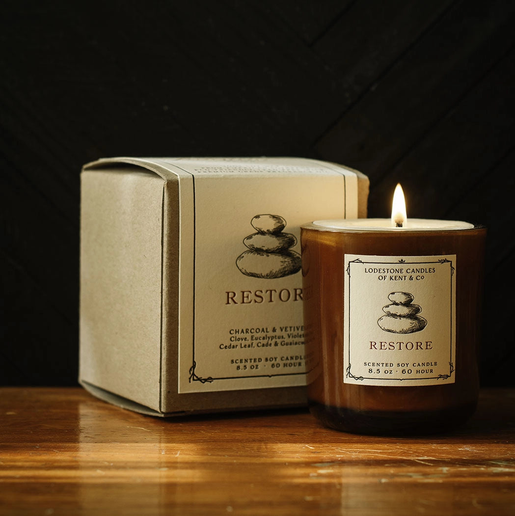 Amber glass jar candle labeled "Restore" with stacked stones icon, lit flame, next to matching cream-colored gift box