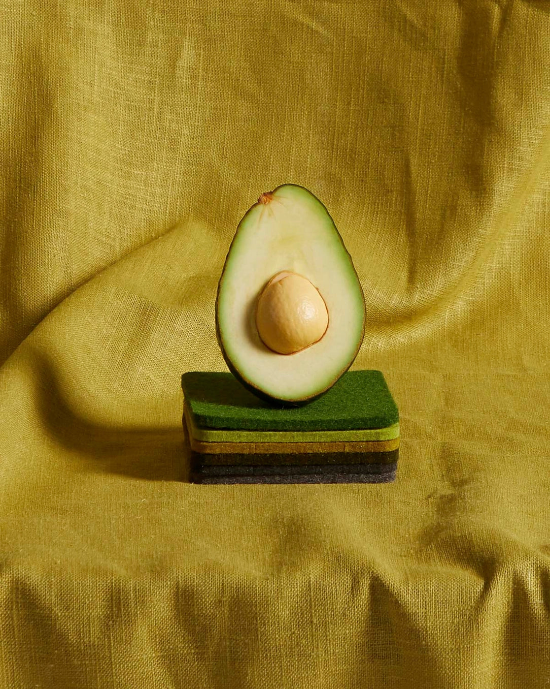 Halved avocado with pit resting on stacked cellulose sponges in earth tones against golden yellow linen backdrop