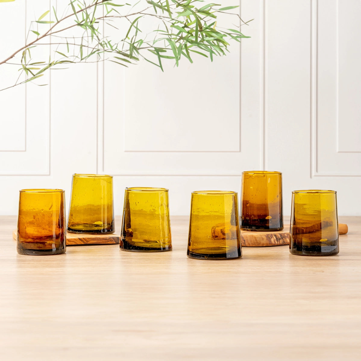 Six amber glass tumblers with tapered shape and bubble texture on wooden surface with white paneled wall background