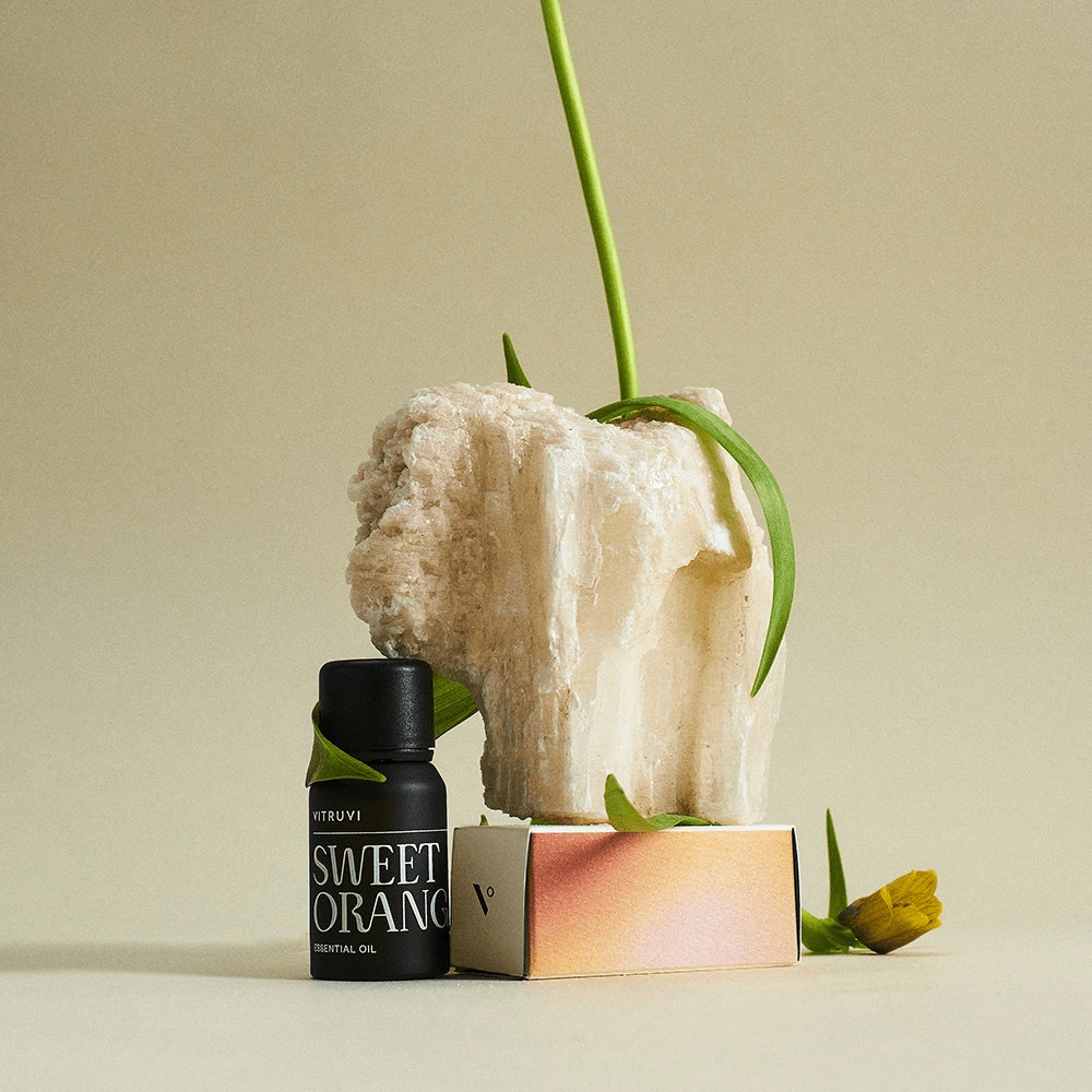 Black essential oil bottle labeled Sweet Orange next to white crystalline rock with air plant and coral-pink gradient box