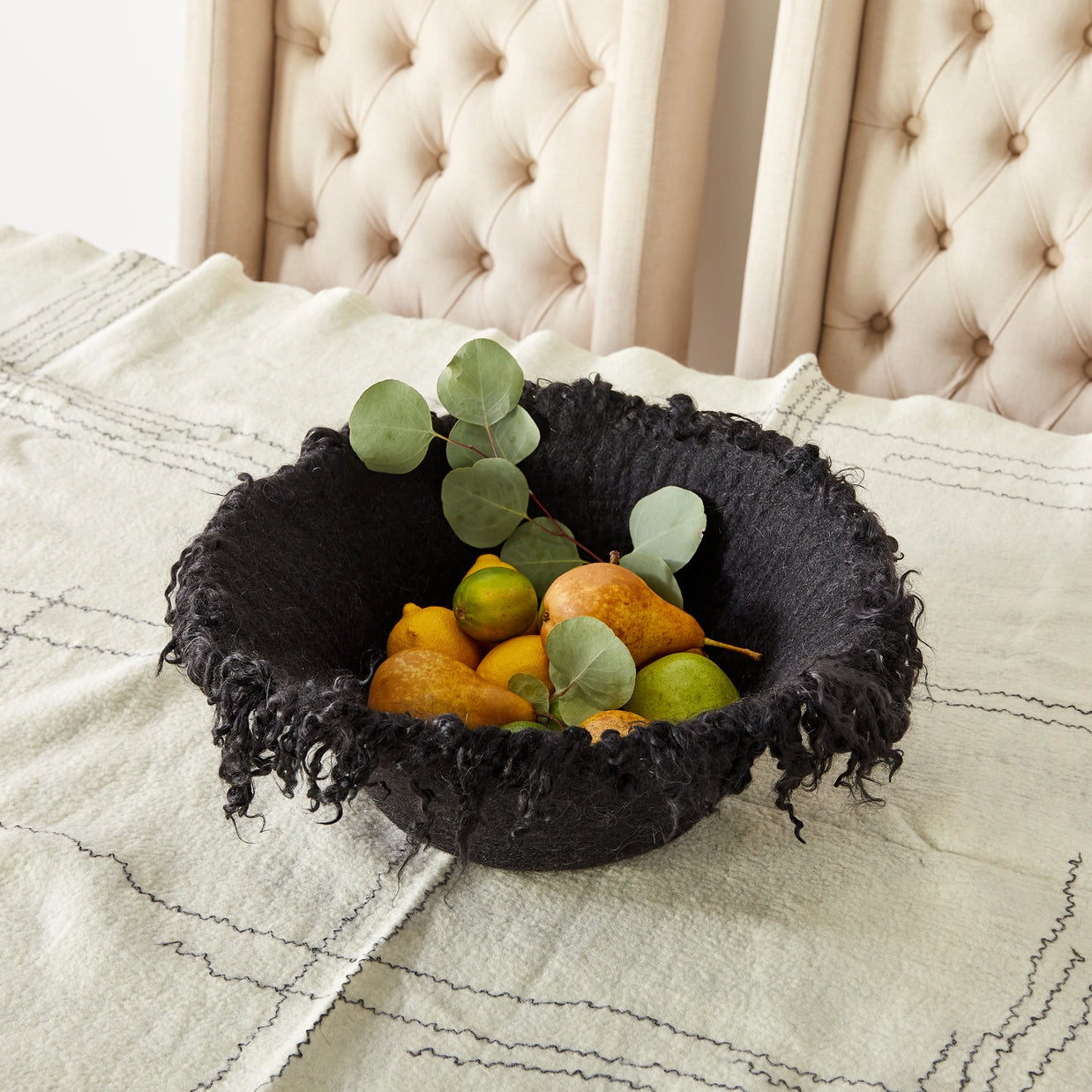 Black woven bowl with frayed textile edges filled with yellow pears, limes, and eucalyptus leaves on cream bedding