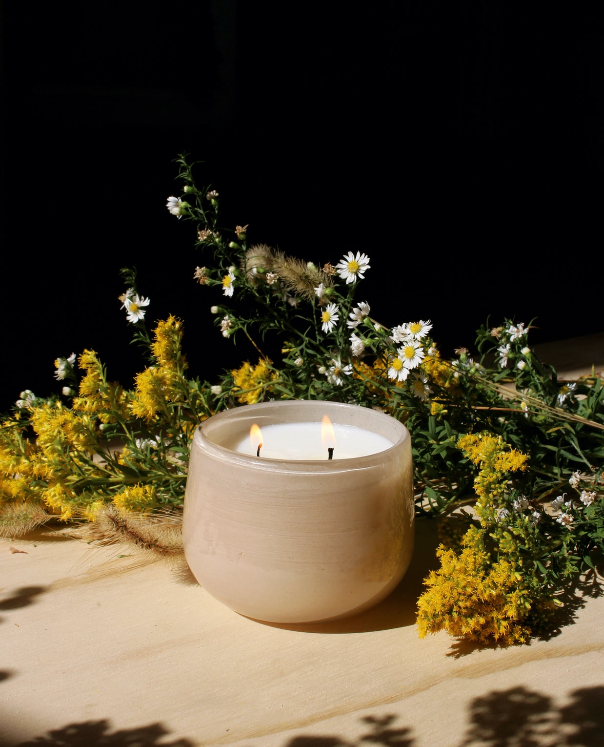 Two-wick candle in beige ceramic vessel with white wax surrounded by yellow goldenrod and white daisy wildflowers