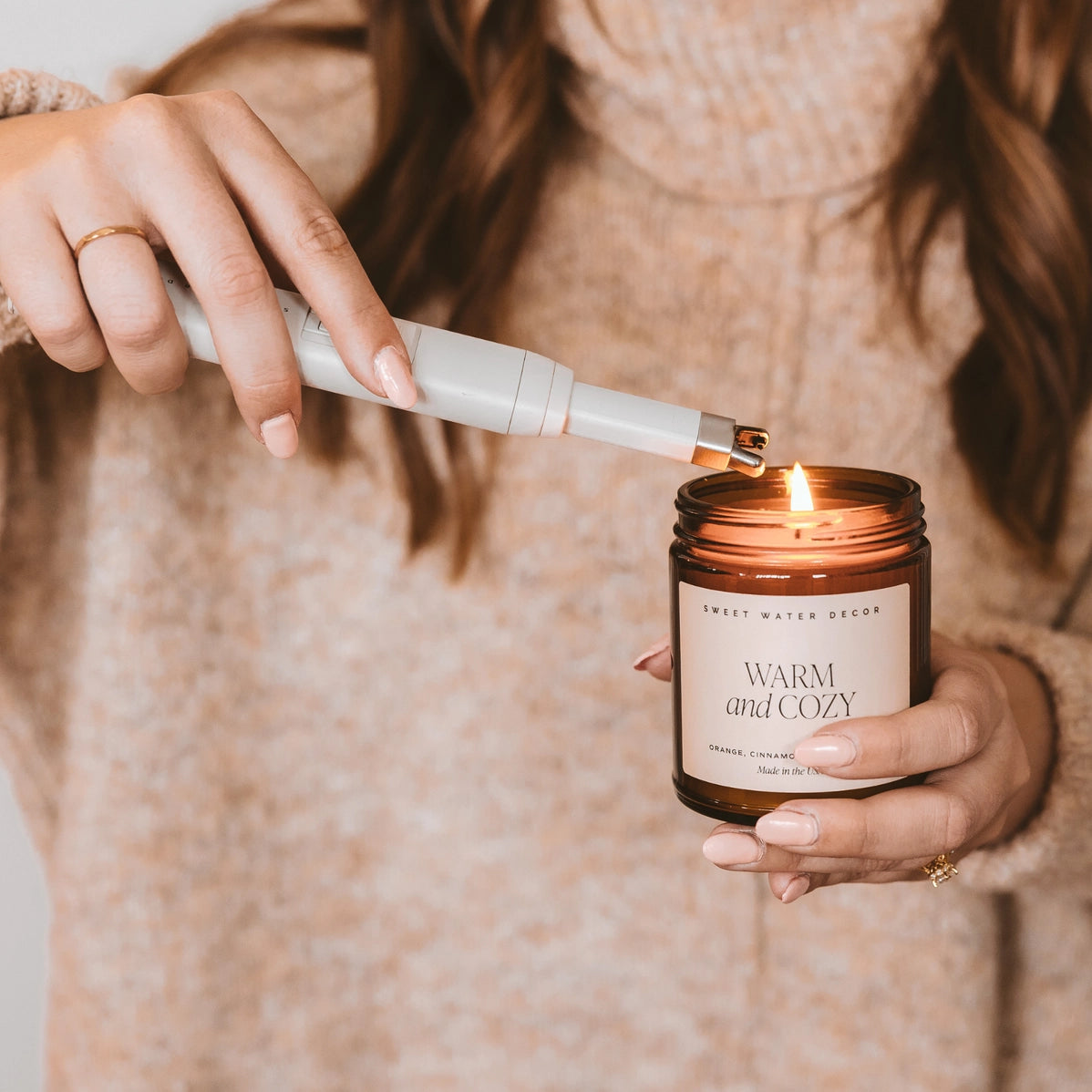 Hand lighting amber glass candle labeled Warm and Cozy with white electric lighter, cream sweater visible in background