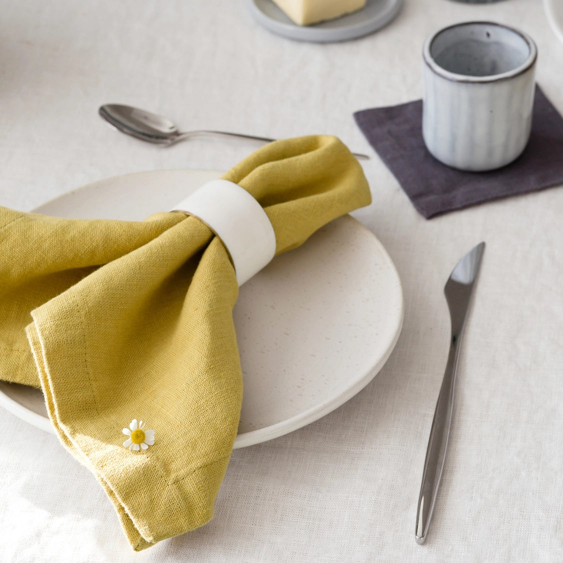 Golden yellow linen napkin with white daisy embroidery held by white ceramic ring on cream stoneware plate with cutlery