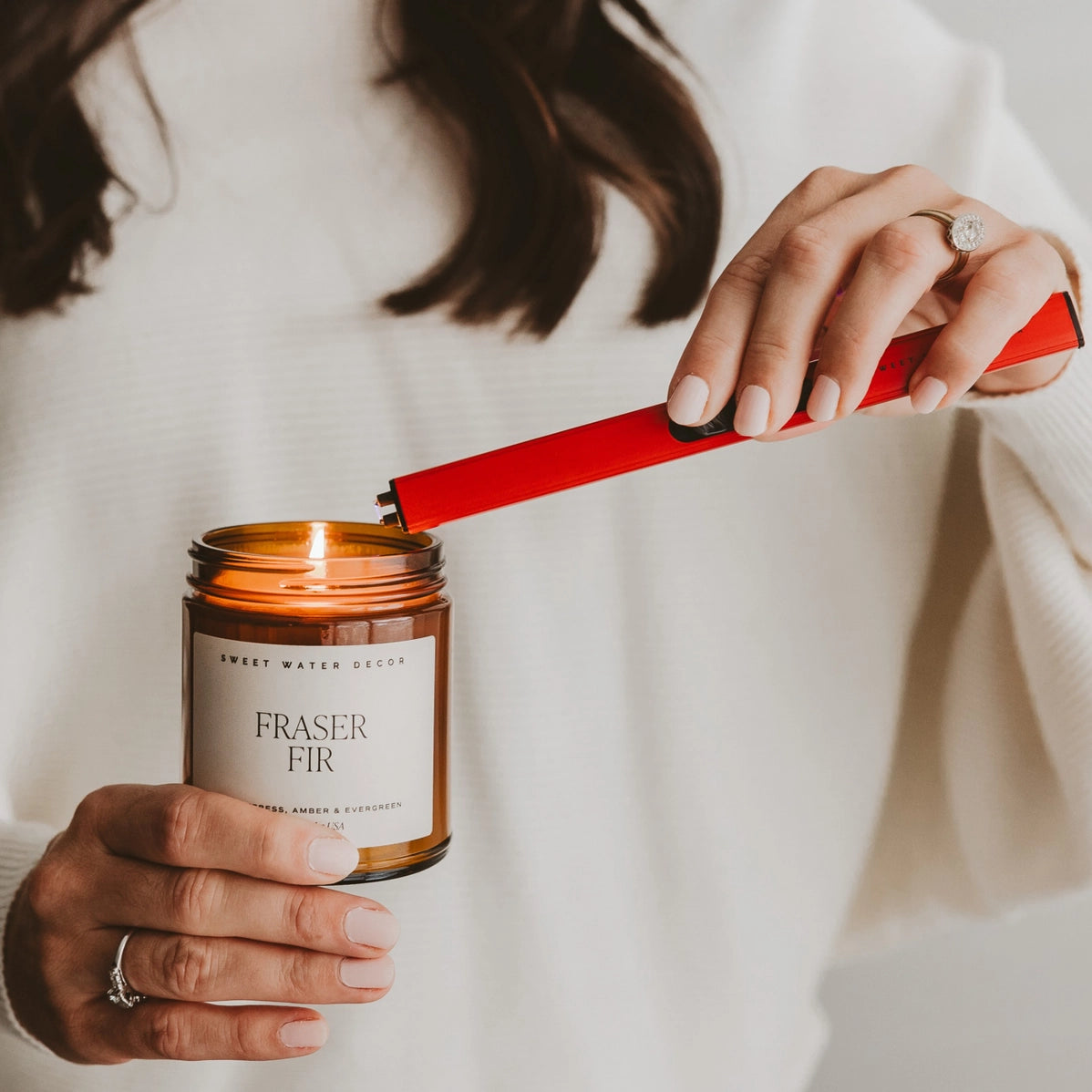 Woman lighting Fraser Fir candle in amber glass jar with bright red match striker, wearing diamond ring on white background
