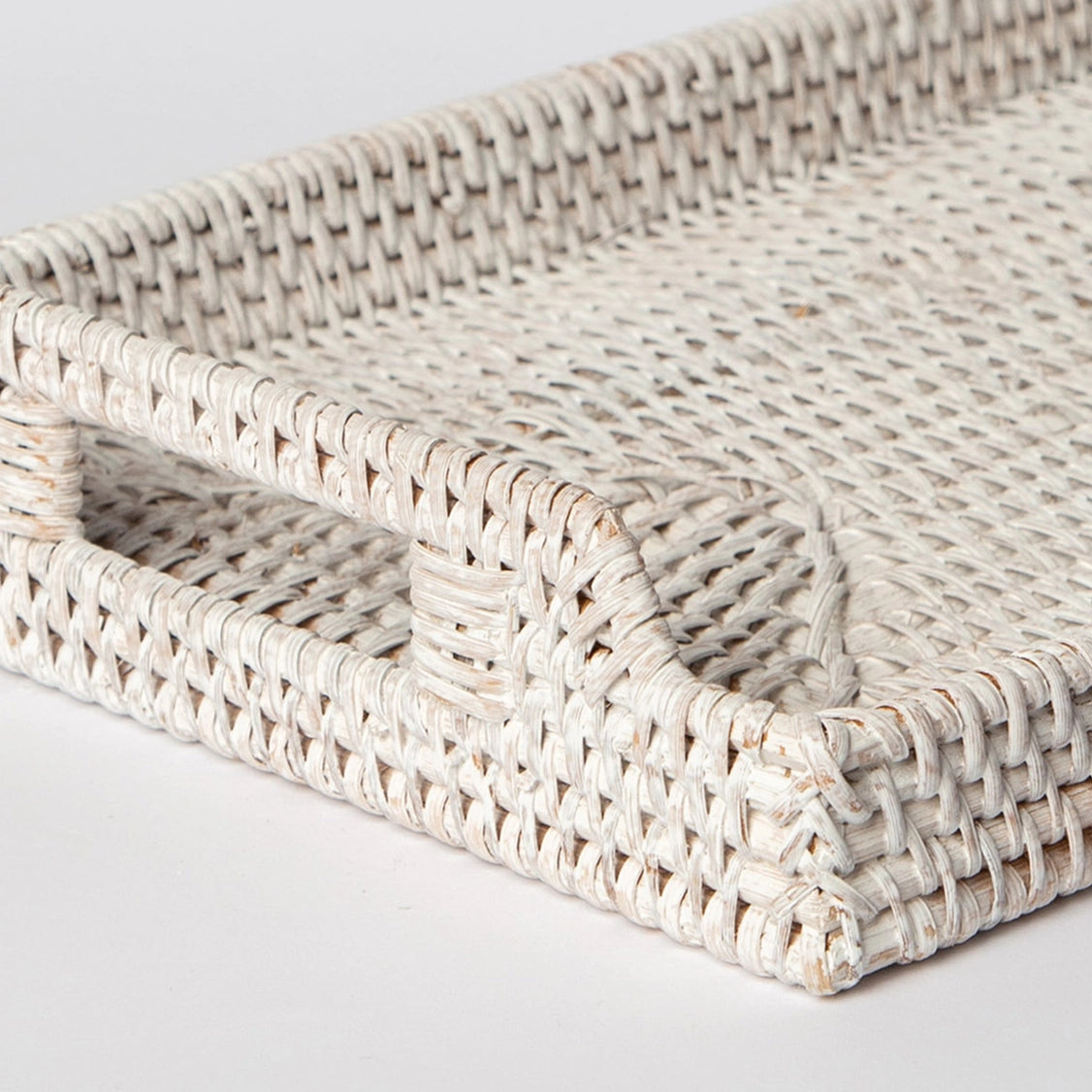 Whitewashed woven rattan storage tray with rounded corners and integrated handles in tight basketweave pattern