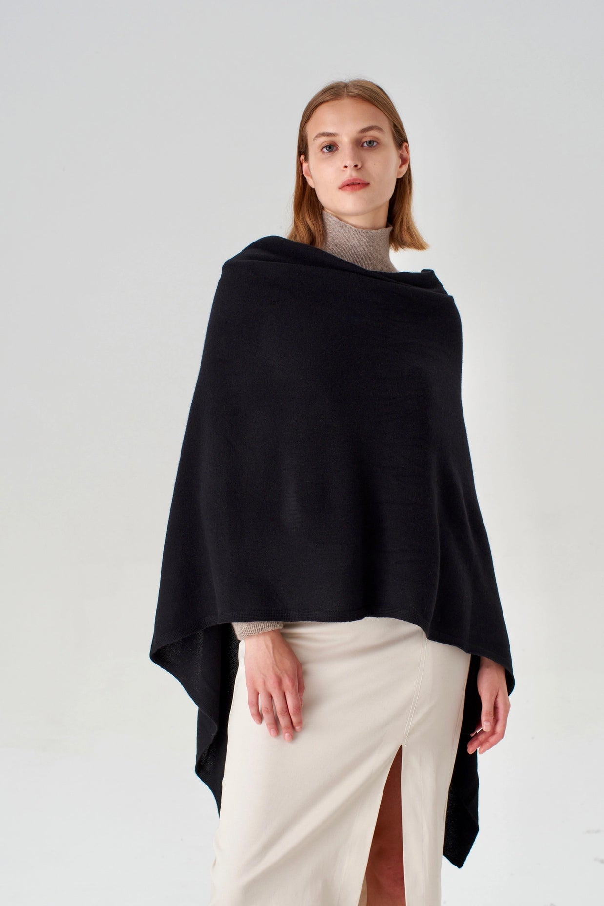 Black knit poncho with asymmetrical hem draped over taupe turtleneck and cream midi skirt with side slit