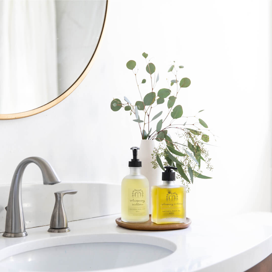 Two frosted glass soap dispensers with black pumps and yellow labels on wooden tray beside eucalyptus branches in white vase