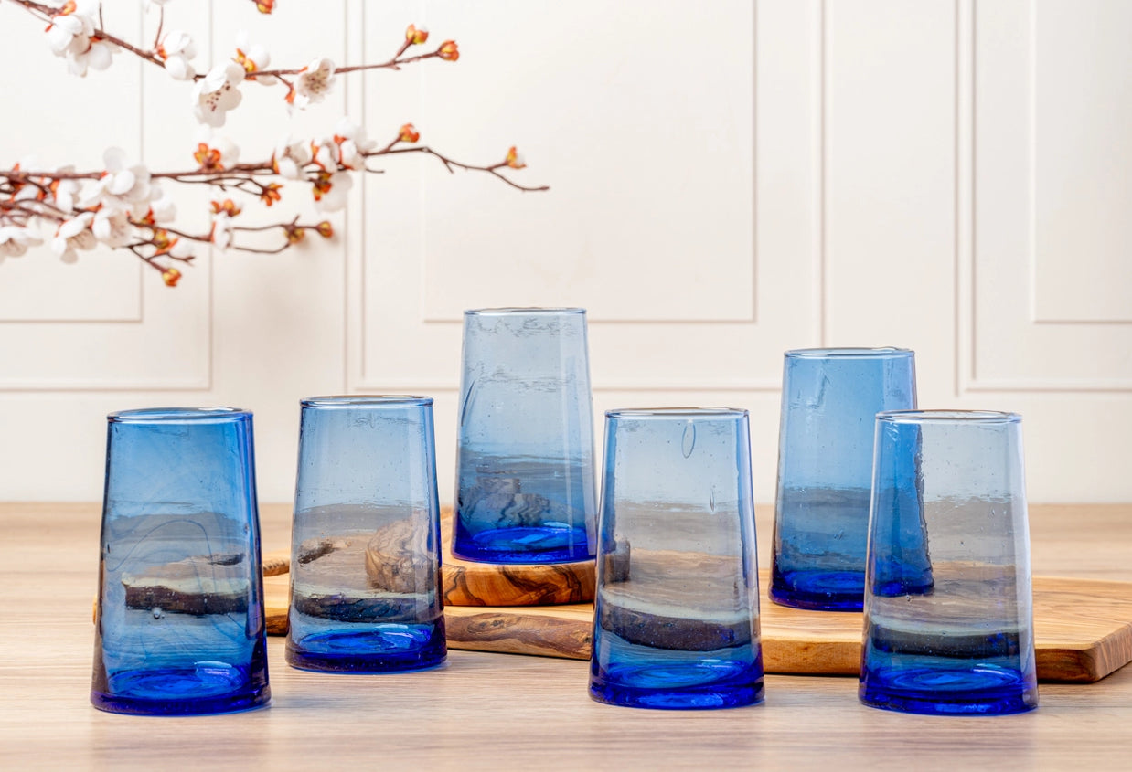 Six hand-blown glass tumblers in gradient blue tones with organic bubbles and natural wood slice bases on light wood surface